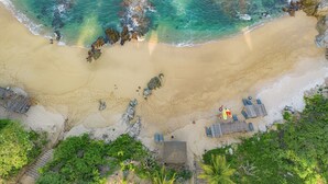 An aerial view of a beach with clear turquoise water, sandy shore, and a few scattered rocks. There are lounge chairs and umbrellas along the beach, and a small building near the water's edge. - Coral Blue Huatulco All Inclusive (Santa María Huatulco)