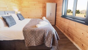 A wooden cabin bedroom with a bed, pillows, a quilted blanket, a wardrobe, and a window with a view of the outdoors. - Bacheiddon Log Cabin (Machynlleth)