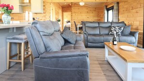 A modern log cabin interior with a grey leather sofa, wooden dining table, and a kitchen area with wooden walls and pendant lighting. - Bacheiddon Log Cabin (Machynlleth)