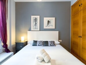 A neatly made bed with two pillows and two rolled towels, a bedside table with a lamp, and two framed artworks on the wall.
