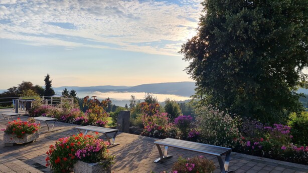 Un parco con panchine, aiuole fiorite e vista su un lago e montagne.