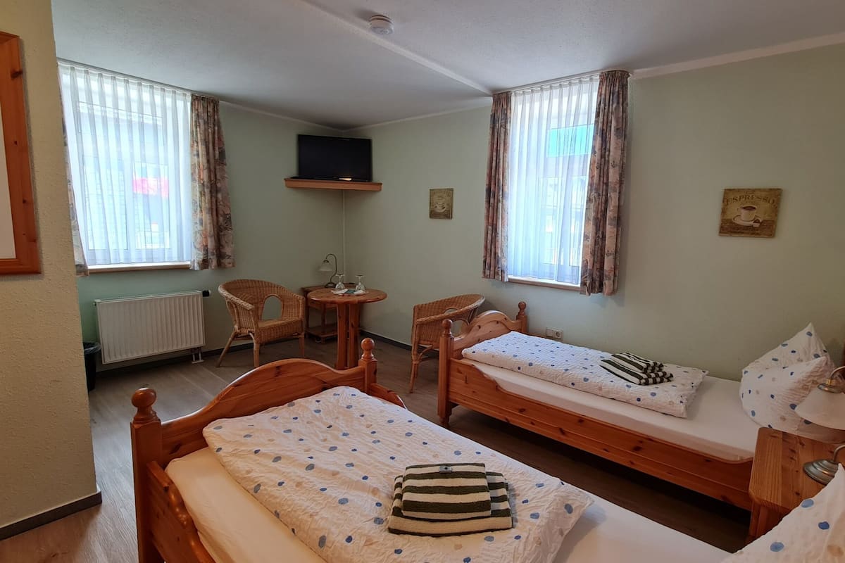 A room with two wooden beds, a chair, a small table, a television, and windows with curtains.