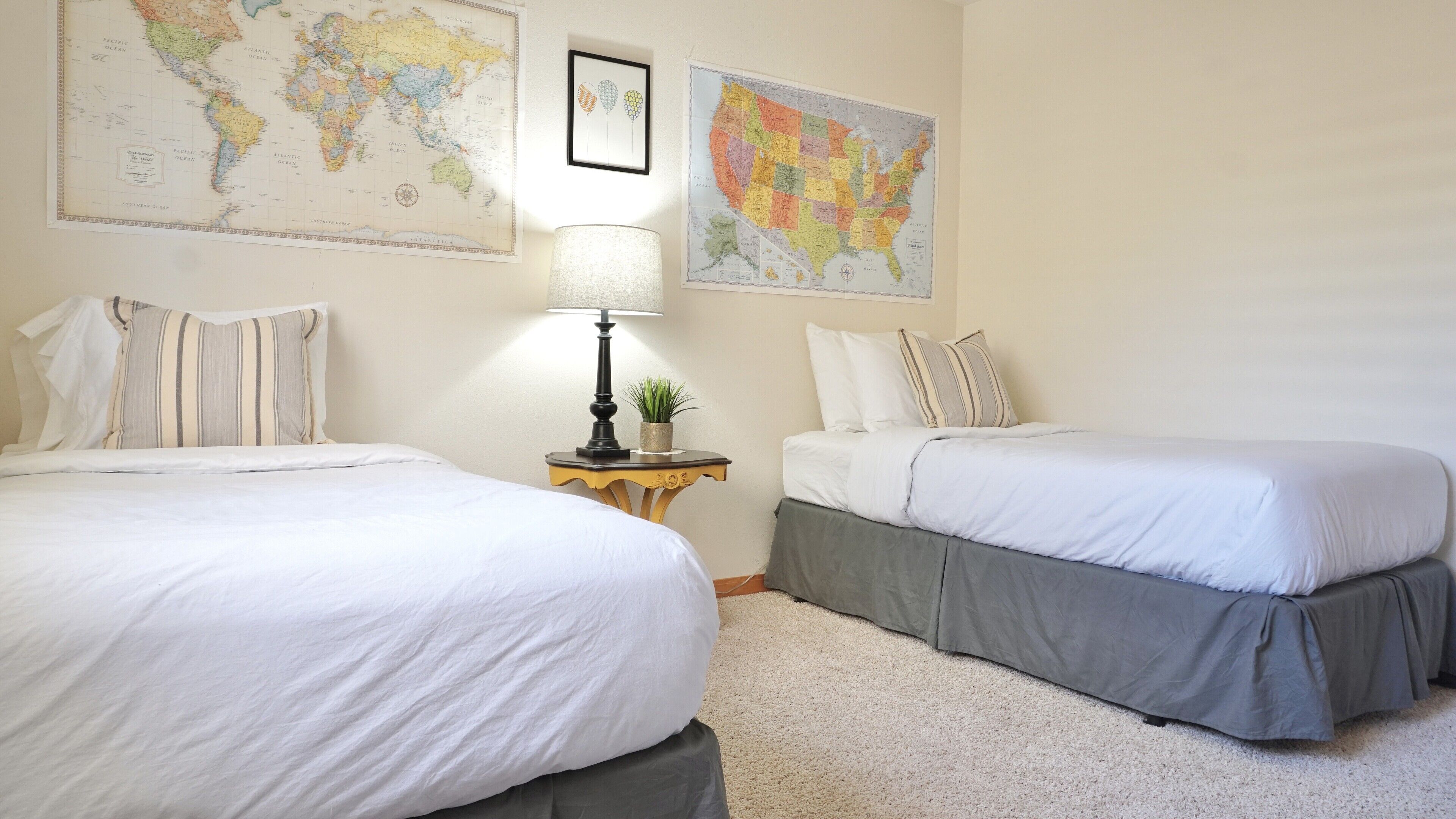 Habitación de hotel con dos camas, una mesita con lámpara y planta, además de dos mapas grandes en la pared.