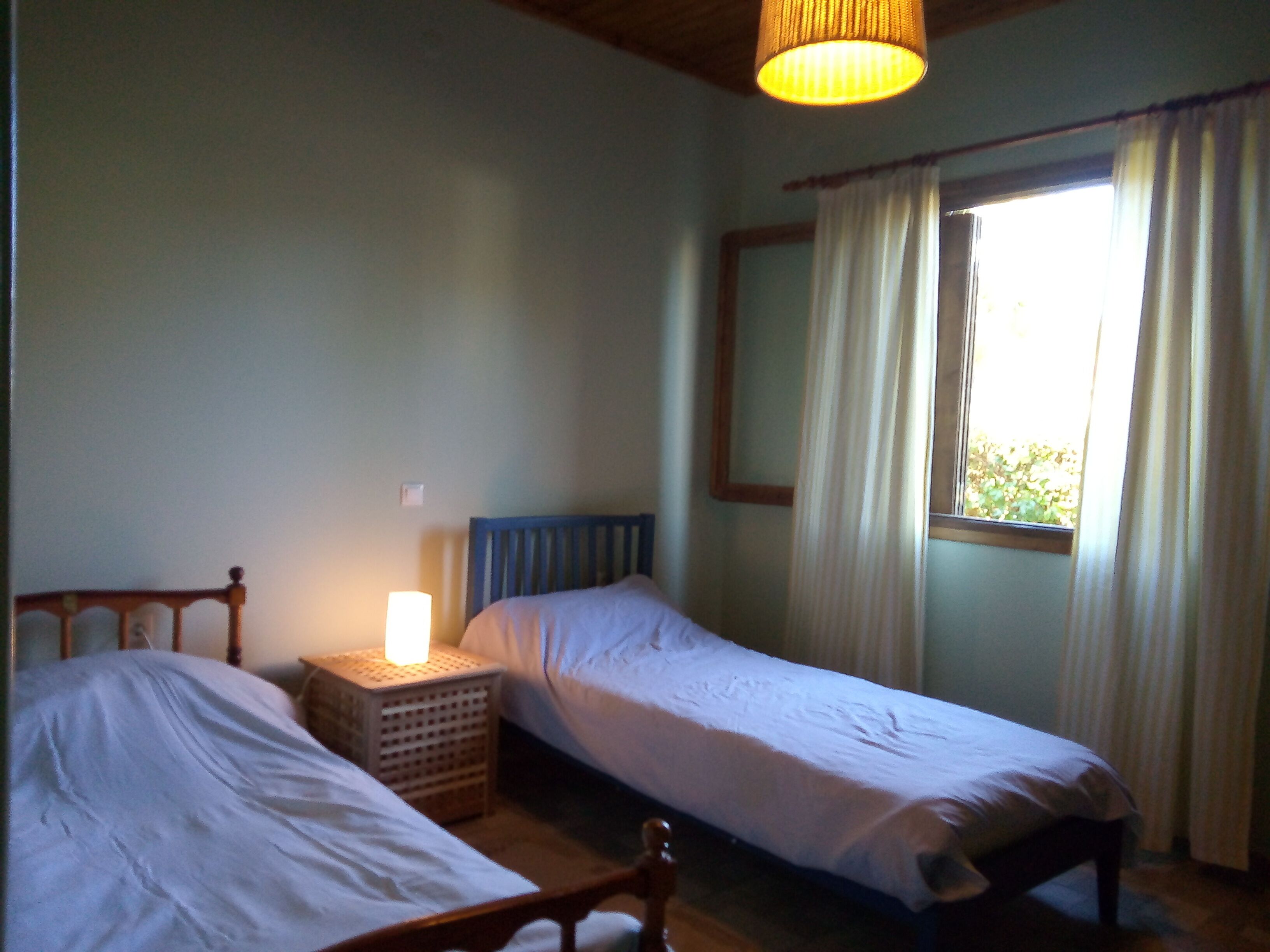 Un dormitorio con dos camas individuales, una mesita de noche con lámpara, una ventana con cortinas y techo de madera.