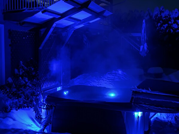 Ein Whirlpool mit blau beleuchtetem Baldachin, umgeben von schneebedeckter Bepflanzung und einer Holzterrasse.