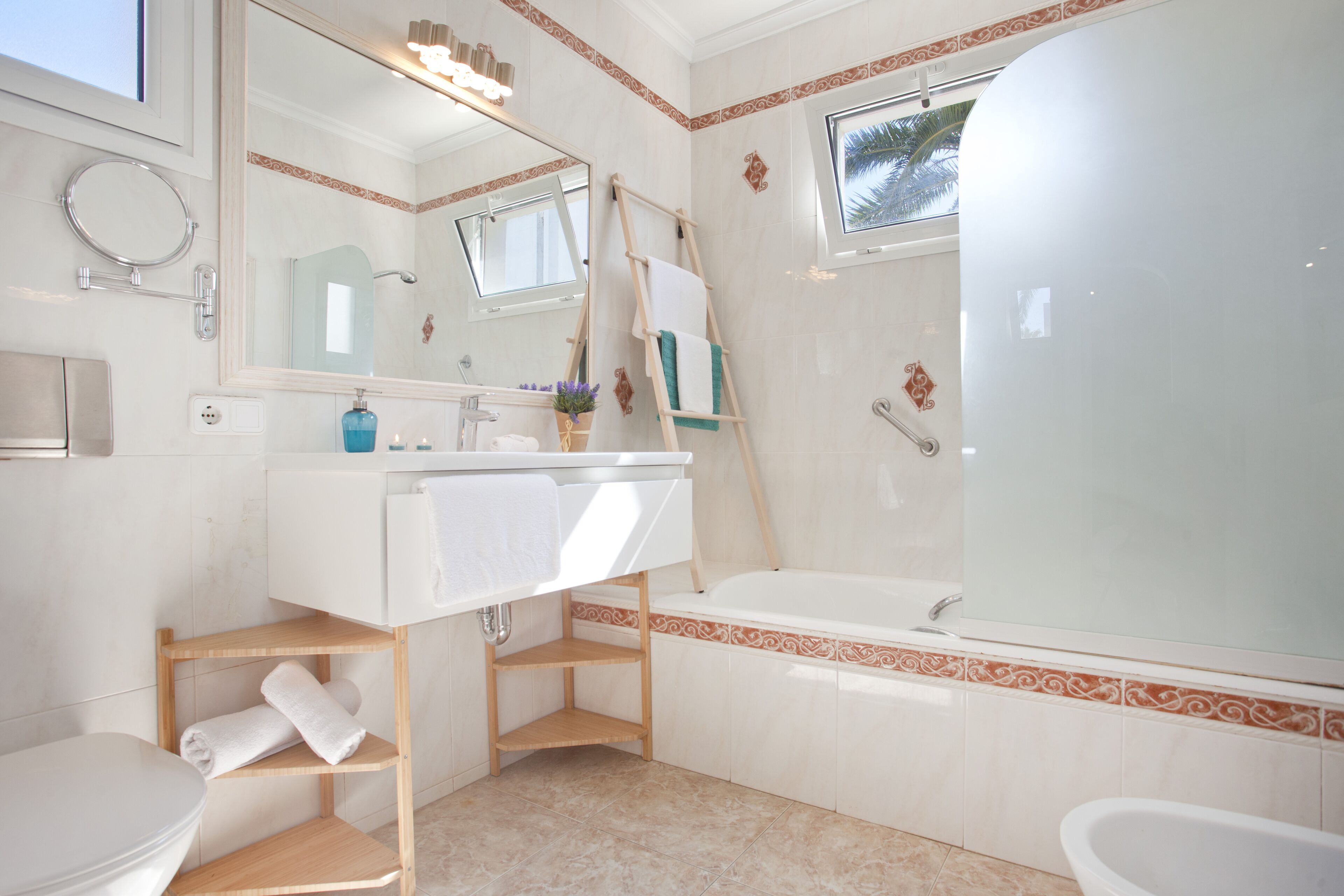 A modern bathroom with a white sink, mirror, and bathtub. There are wooden shelves under the sink and a window above the bathtub.