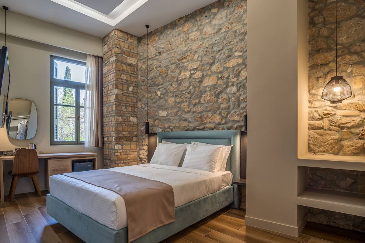 A bedroom with a stone wall, a bed with white bedding, a desk with a chair, and a hanging light fixture.