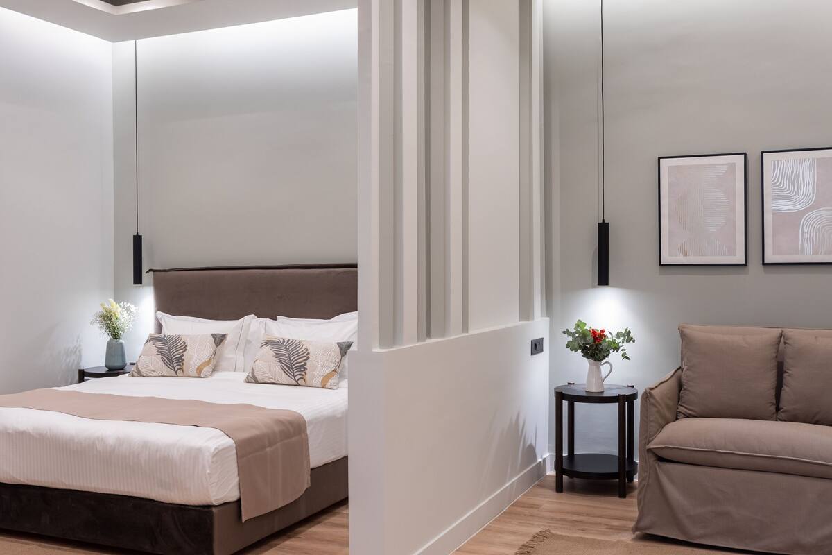 A modern bedroom with a large bed, a sofa, a small table with a vase, and a wall-mounted shelf.