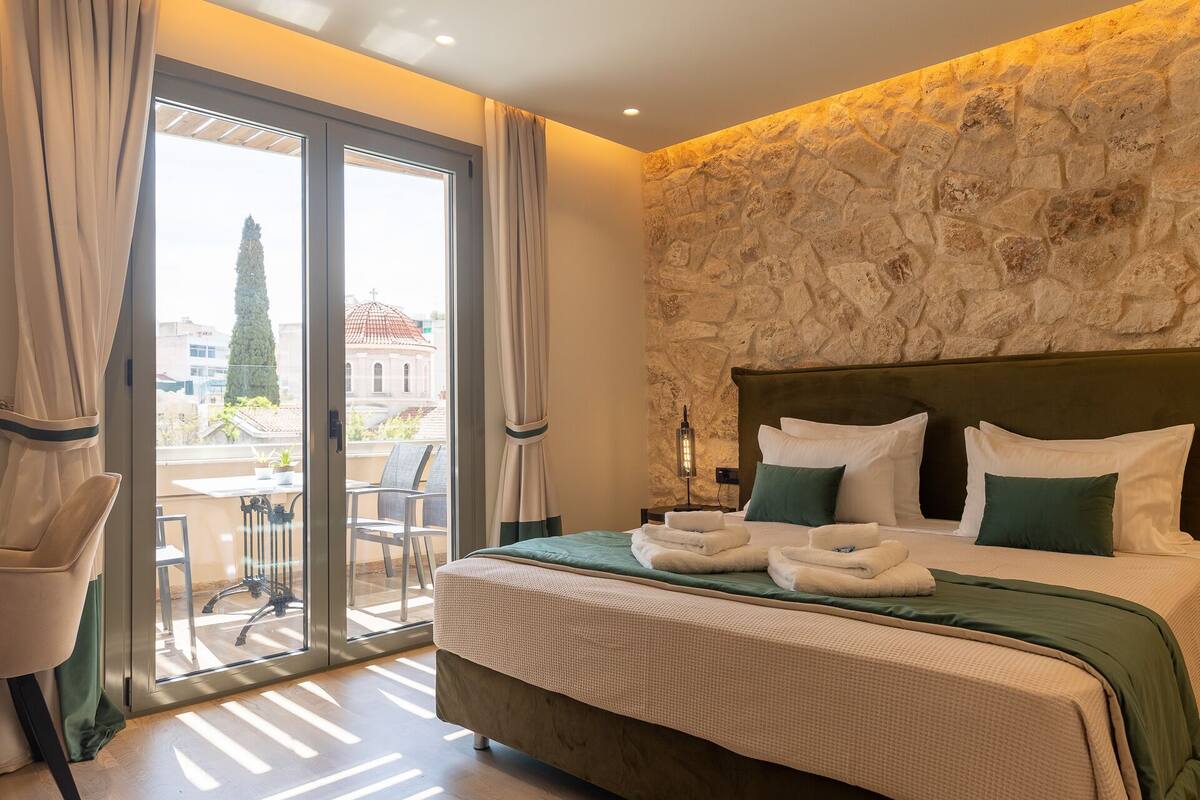 A hotel room with a bed, a view of the city, and a balcony with chairs.