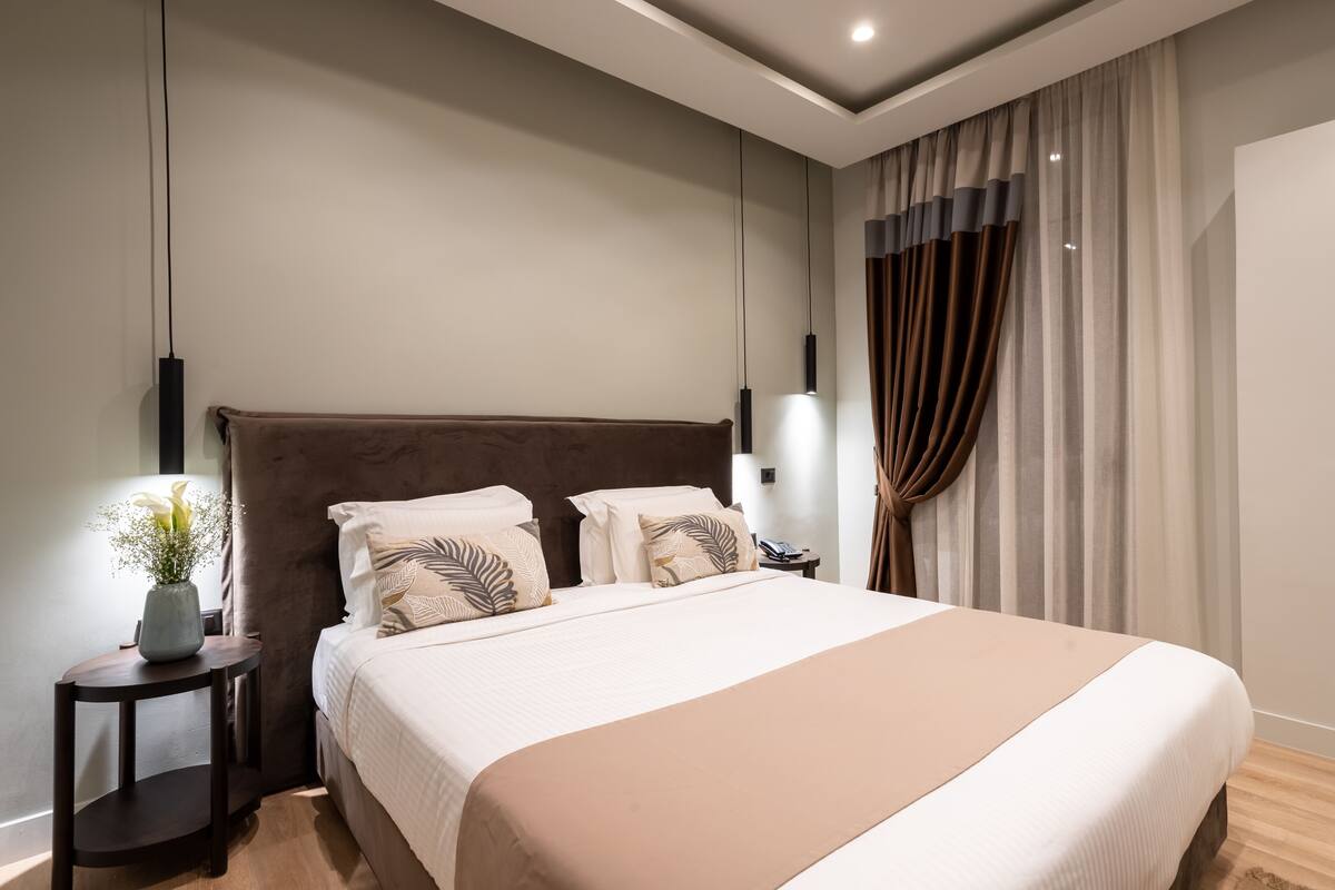 A modern hotel room with a large bed, a bedside table with a vase of flowers, and a view of the city through the window.