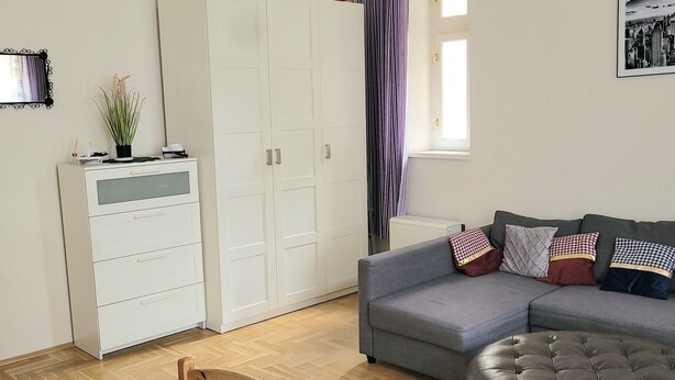 A bedroom with a bed, a grey sofa, a wooden bench, a white wardrobe, and a window with curtains. - Apartament Rachel Stare Miasto Basztowa 23 (Kraków)