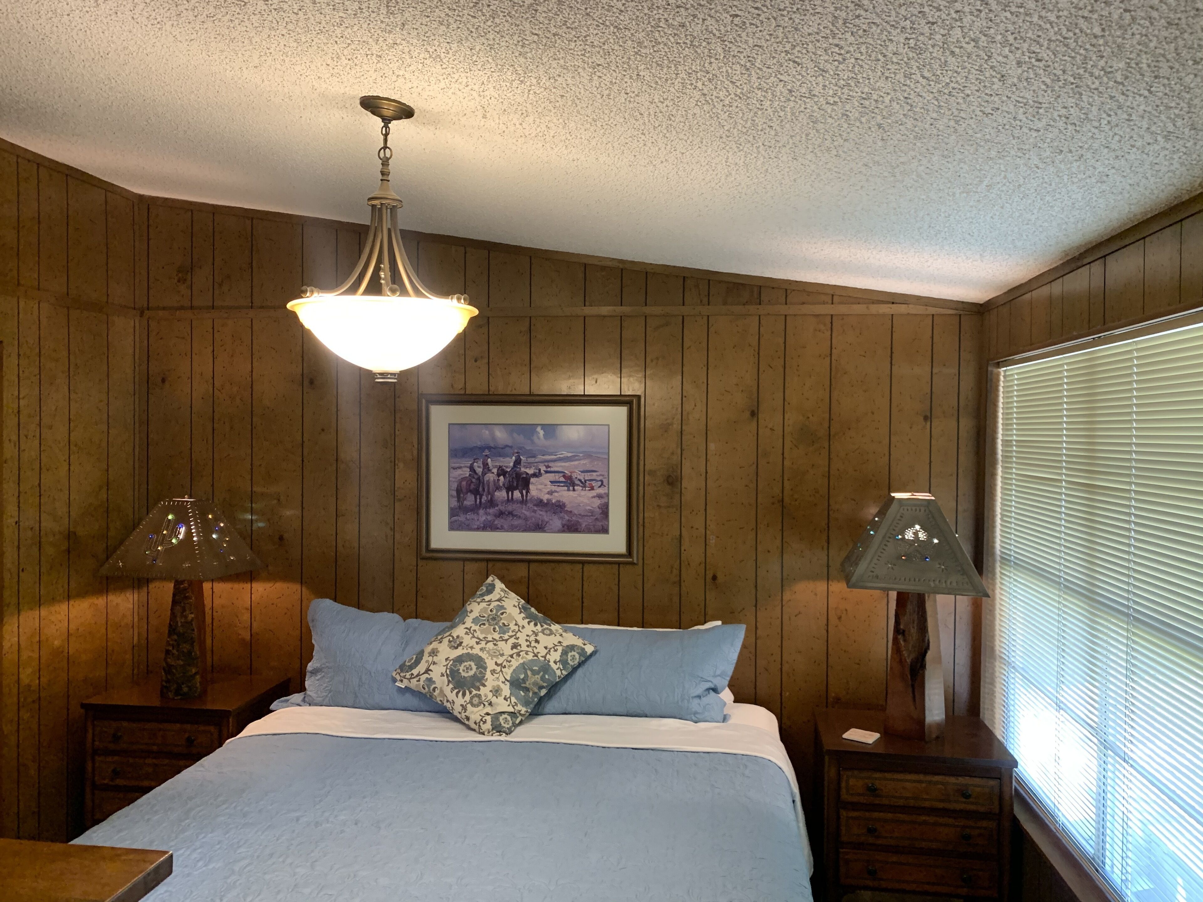 A bedroom with wooden paneling, a bed with blue bedding, a framed picture on the wall, and a window with blinds.