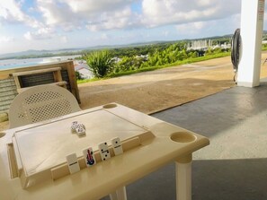 A table with dominoes and a view of a landscape with greenery and a body of water. - Ocean View Sunset (Fajardo)