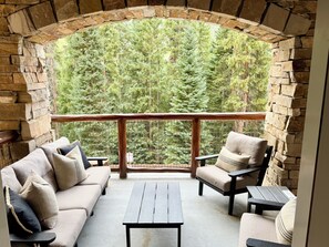 A stone-walled balcony with a view of dense forest, furnished with a sofa, armchair, and a small table.