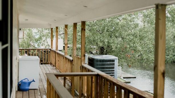Une terrasse en bois avec une rambarde, un climatiseur blanc et un arrosoir bleu.