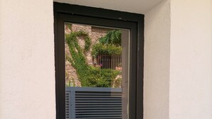 Une petite fenêtre à cadre noir, un pot de fleurs et une vue sur un bâtiment en pierre, agrémenté de verdure.