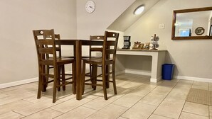 Un coin repas avec une table ronde et quatre chaises, une horloge au mur, un bureau avec un ordinateur et une machine à café, ainsi qu’une poubelle bleue.