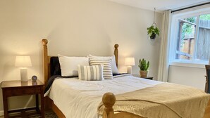 Un lit en bois avec une literie blanche, deux tables de chevet avec des lampes, une plante en pot et une fenêtre donnant sur une clôture en bois.