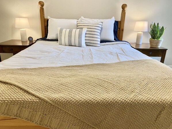 A neatly made bed with a knitted blanket, two bedside lamps, a potted plant, and a wooden headboard.