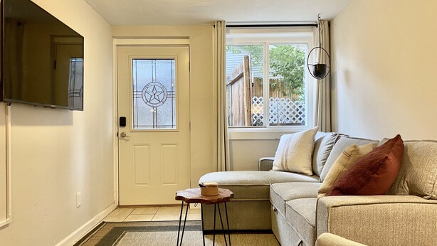 Un salon avec un canapé beige, une petite table en bois, un téléviseur fixé au mur et une fenêtre donnant sur une clôture en bois et des arbres.