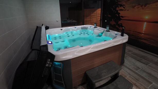 A modern bathroom with a large, illuminated hot tub, a wooden bench, and a view of a sunset through a large window. - Wellness in Sologne La Frenaudiere spa (2 Villa la fronodiere. Gy-en-Sologne)
