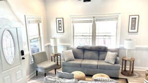 A living room with a grey sofa, two armchairs, a coffee table, and a ceiling fan. There are two windows with blinds, a door with a glass panel, and a small side table with a lamp. - Beautiful Beach Home -3 min walk to beach -with golf cart option (Myrtle Beach)