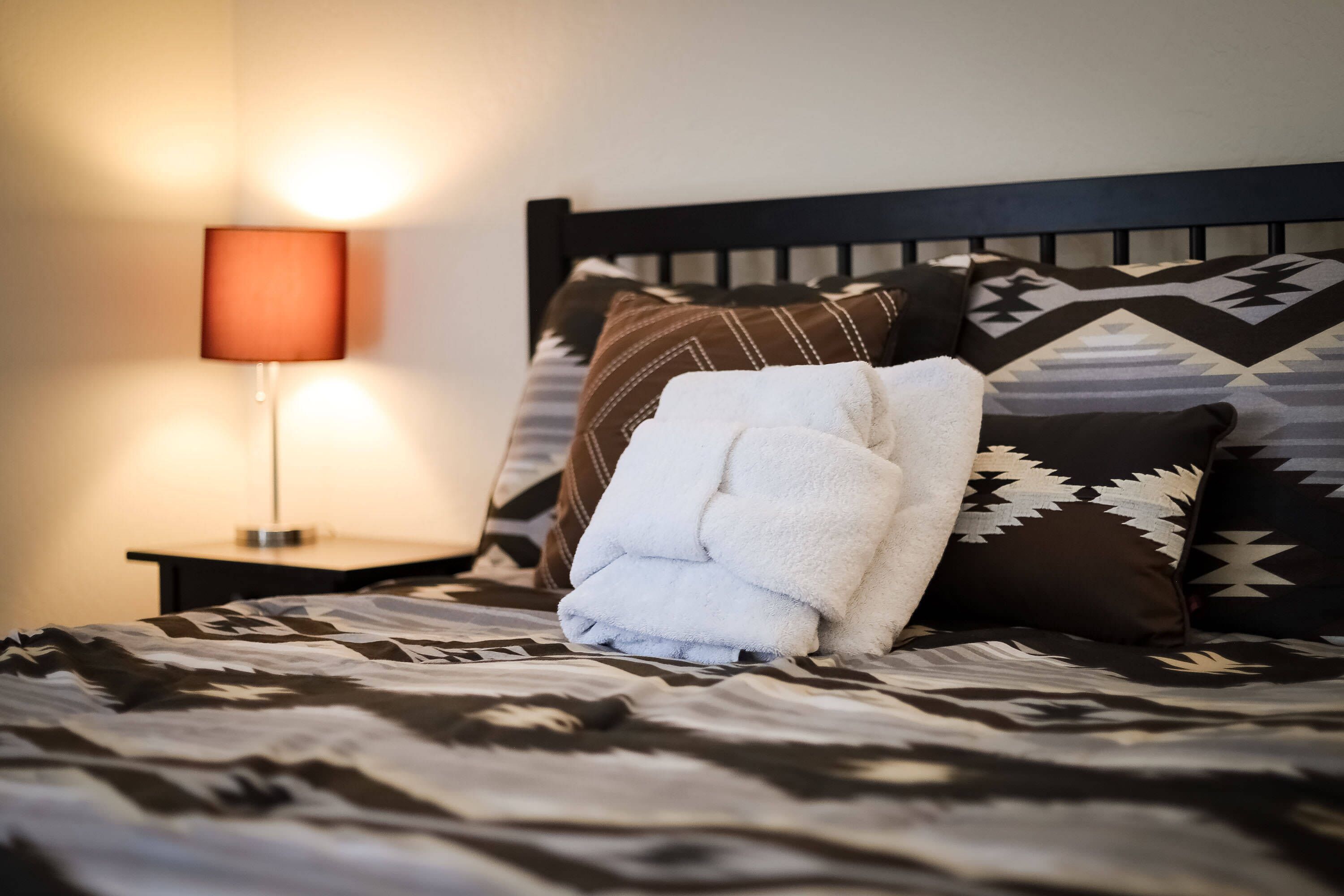 Ein ordentlich bezogenes Bett mit einer gemusterten Tagesdecke, einem gefalteten weißen Handtuch und dekorativen Kissen auf einem dunklen Kopfteil.
