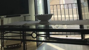 Una habitación con una televisión sobre un soporte, una mesa de vidrio con un cuenco blanco, y un balcón con barandilla y una escalera.