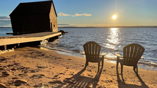 Une jetée en bois s’avance sur des eaux calmes, et une petite construction se trouve à son extrémité. Deux chaises Adirondack sont placées sur la plage de sable, face à l’eau.