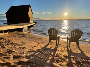 Een houten pier die zich uitstrekt over kalm water, met een klein gebouw aan het einde. Twee Adirondack-stoelen staan op het zandstrand, met uitzicht op het water.