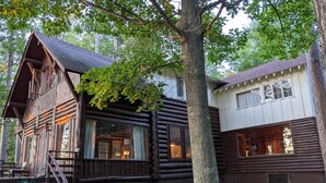 Une cabane rustique en rondins de bois, avec un escalier en bois menant à un porche, le tout entouré d’une végétation luxuriante.