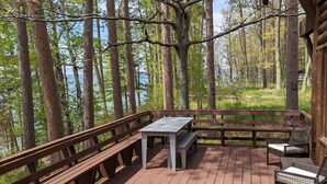Une terrasse en bois avec une table et des bancs, entourée de grands arbres et offrant une vue sur une étendue d’eau.