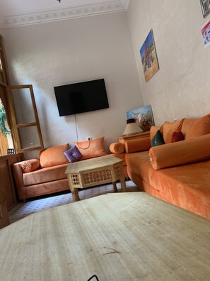 A living room with two orange sofas, a wooden coffee table, a TV mounted on the wall, and a painting. - RIADJENANADAM Marrakesh (Marrakech)