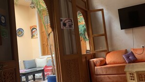 A room with a wooden door, a sofa with orange cushions, a TV mounted on the wall, and a patterned floor. - RIADJENANADAM Marrakesh  (Marrakech)