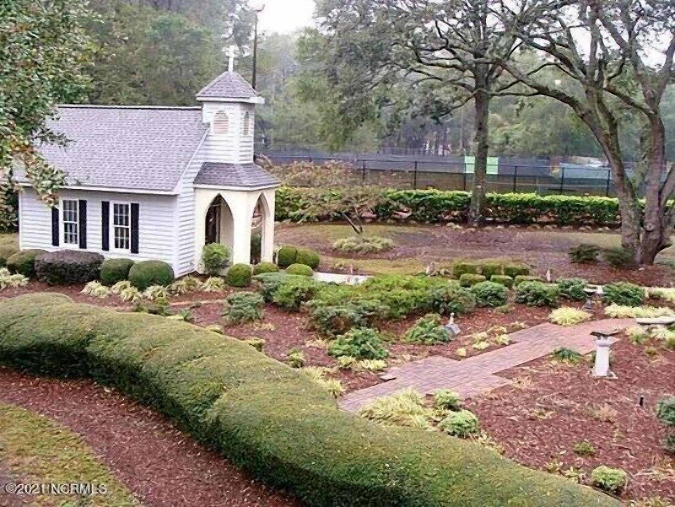 A small white church with a steeple, surrounded by well-manicured hedges and a brick pathway.