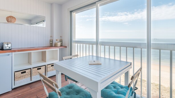 A room with a white table and chairs, a wooden floor, a large window with a view of the beach, and a white ceiling. - LE PANORAMIQUE - Face mer - Wifi (Pornichet)