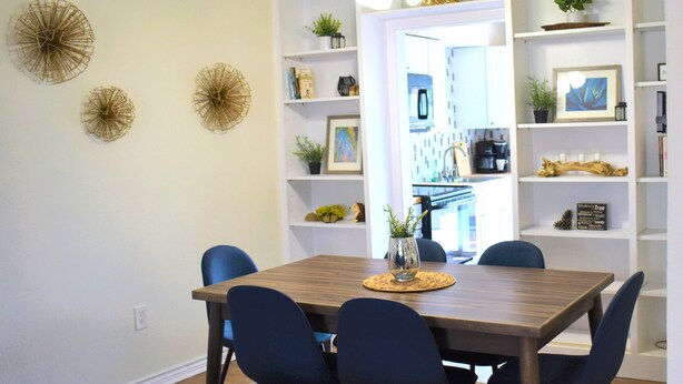 A dining area with a wooden table, four chairs, decorative wall art, and built-in shelving with plants and books. - Feel at Home at this central and spacious 1 bedroom condo (Dallas)
