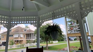 A covered porch with a ceiling fan, a hanging planter, and a view of a residential area with houses and a white picket fence.