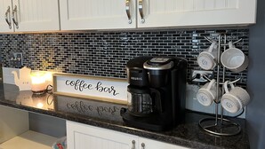A kitchen with white cabinets, a coffee maker, a mug rack, and a sign that reads "coffee bar".