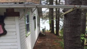 Un étroit sentier mène à une maison blanche aux bordures vertes, entourée d’arbres.