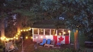 Un espace extérieur aménagé avec une zone de sièges couverte, décoré aux couleurs du drapeau canadien, comprenant une petite terrasse et des guirlandes lumineuses.