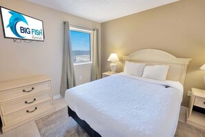 Una habitación de hotel con una cama grande, una mesita de noche, un aparador, un televisor mostrando 'BIG FISH Rentals' y una ventana con cortinas.