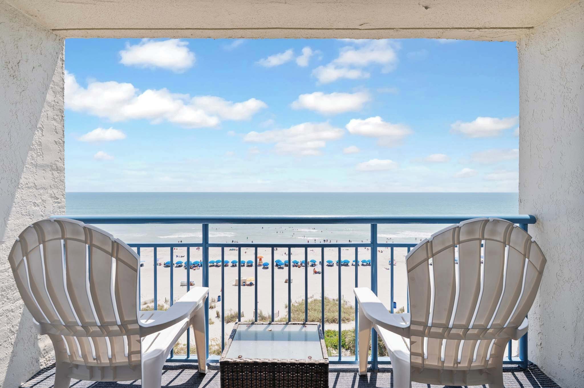 A balcony with two Adirondack chairs facing a beach with umbrellas and people.