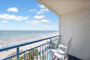 A balcony with a view of a beach, a pool, and a clear sky.