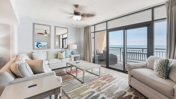 A spacious living room with a large sliding glass door leading to a balcony, a ceiling fan, and a view of the ocean. - Ocean-view condo with balcony & beach access (Myrtle Beach)