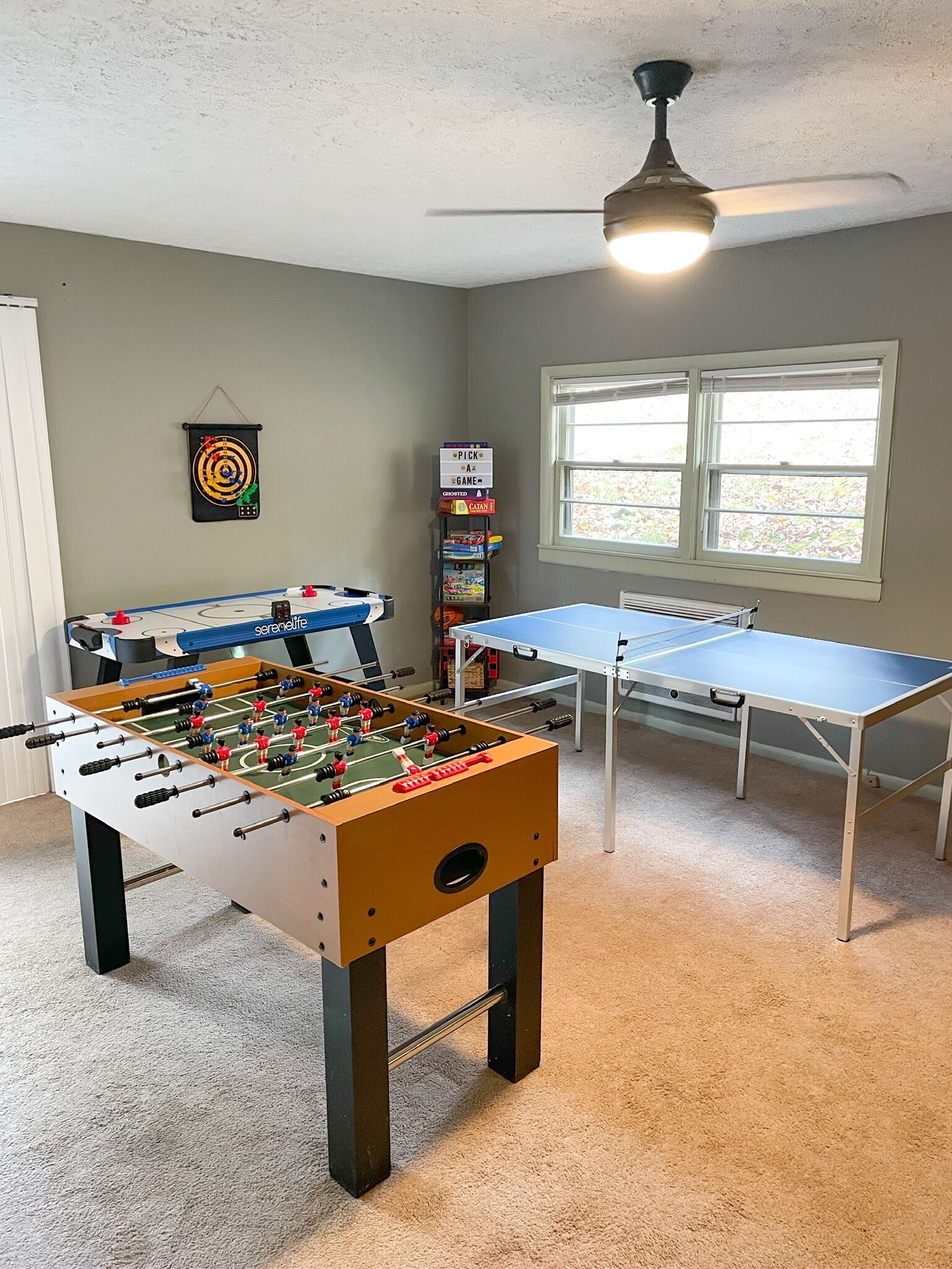 A room with a foosball table, a ping pong table, and a dartboard. There is a window with blinds and a ceiling fan.