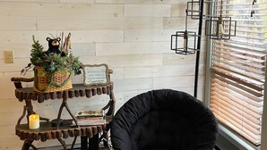 A room with a wooden floor, a black chair, a wooden wall, a window with blinds, and a decorative wooden shelf with a basket and a sign. - Spacious 3BR/3BA Cabin Only 0.5 Miles From Slopes! (Beech Mountain)