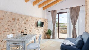Una habitación con pared de piedra rústica, techo de madera, televisor montado en la pared, una mesa con sillas, un sofá y un amplio ventanal con cortinas.