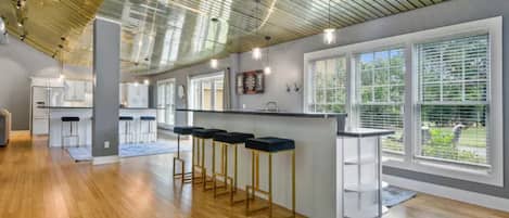 A modern kitchen with a bar area, large windows, and a high ceiling with exposed beams.