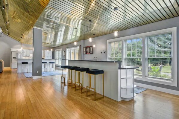 A modern kitchen with a bar area, large windows, and a high ceiling with exposed beams.